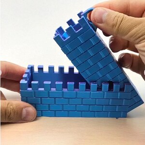 May include: A blue toy castle with a hinged lid, constructed from brick-like blocks. The lid is being lifted, revealing the interior. The toy is designed with crenellations along the top edges.