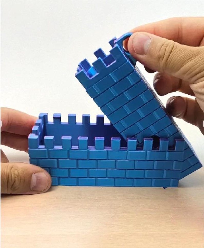 May include: A blue toy castle with a hinged lid, constructed from brick-like blocks. The lid is being lifted, revealing the interior. The toy is designed with crenellations along the top edges.