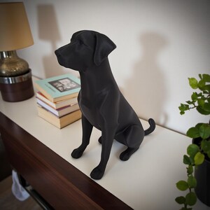 K&ouml;nnte beinhalten: Eine matte, schwarze Labrador Retriever-Statue in sitzender Position. Die Hundefigur steht auf einer wei&szlig;en Oberfl&auml;che, im Hintergrund sind B&uuml;cher und eine Lampe zu sehen. Der Schwanz des Hundes ist eingerollt.