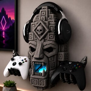 May include: A gray stone-textured wall mount shaped like a carved totem head with a skull emblem, featuring two claw-like hands holding a white and a black gaming controller, with black over-ear headphones resting on top and a glowing blue light.