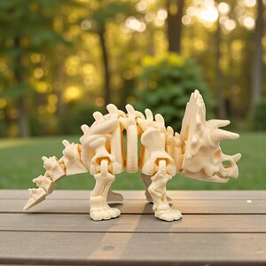 Może przedstawiać: Beżowy, przegubowy model szkieletu dinozaura Triceratops. Model wykonany jest z jasnego materiału i ustawiony na drewnianej powierzchni. Dinozaur ma kołnierz, rogi i segmentowy ogon.