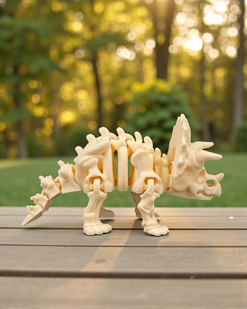 Może przedstawiać: Beżowy, przegubowy model szkieletu dinozaura Triceratops. Model wykonany jest z jasnego materiału i ustawiony na drewnianej powierzchni. Dinozaur ma kołnierz, rogi i segmentowy ogon.