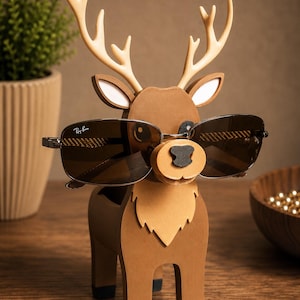 May include: A brown wooden deer-shaped glasses holder with a pair of sunglasses. The deer has tan antlers, white-lined ears, and a black nose. The sunglasses are dark-tinted with a silver frame. The holder is on a wooden surface.