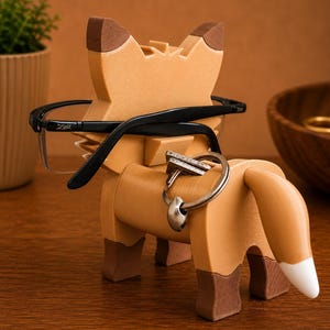 May include: A wooden fox-shaped eyeglasses holder in shades of brown. The fox has black eyeglasses resting on its head and a silver keyring with a ring on its back. The fox's tail has a white tip. The fox is on a wooden surface.