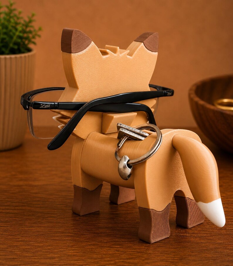 May include: A wooden fox-shaped eyeglasses holder in shades of brown. The fox has black eyeglasses resting on its head and a silver keyring with a ring on its back. The fox's tail has a white tip. The fox is on a wooden surface.