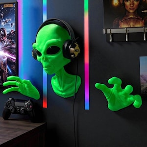 Puede incluir: Decoración de pared con cabeza de alienígena verde con auriculares negros y dos manos verdes. El alienígena está montado en una pared negra. Un mando de juegos negro está sobre una superficie de madera.