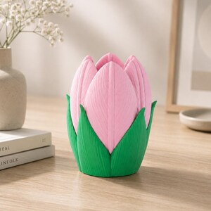 May include: A pink tulip-shaped ornament with green leaves. The object is on a wooden surface. A vase of white flowers is in the background.