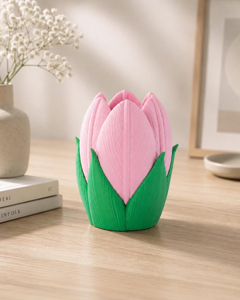 May include: A pink tulip-shaped ornament with green leaves. The object is on a wooden surface. A vase of white flowers is in the background.
