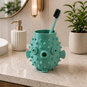 May include: A turquoise toothbrush holder shaped like a pufferfish, with a matching toothbrush. The holder has a textured surface with protruding spikes, eye-like circles, and fin details. The toothbrush has a black bristle head. Bathroom decor.