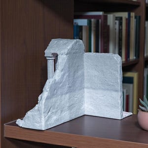 May include: A gray, stone-like bookend shaped like a classical ruin. The bookend features a column and a partially collapsed wall, creating a unique architectural accent. It is placed on a dark brown shelf in front of a bookshelf.