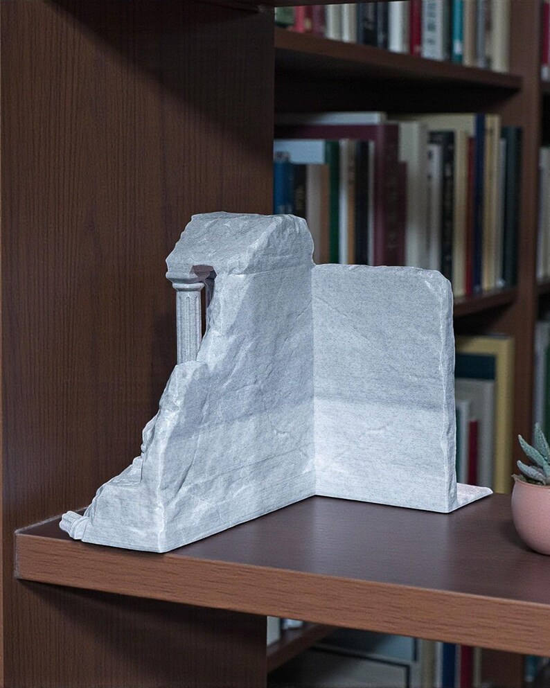 May include: A gray, stone-like bookend shaped like a classical ruin. The bookend features a column and a partially collapsed wall, creating a unique architectural accent. It is placed on a dark brown shelf in front of a bookshelf.
