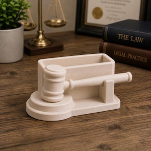 May include: A light-colored wooden gavel and desk organizer. The gavel is resting on a tiered base, with a rectangular organizer behind it. The desk organizer has the words "THE LAW" and "LEGAL PRACTICE" on the spines of the books in the background.