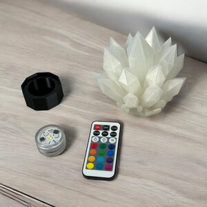 May include: A white crystal-shaped LED light with a remote control, a black cylindrical holder, and a small silver LED light. The remote has color options. The items are on a light wood-grain surface.