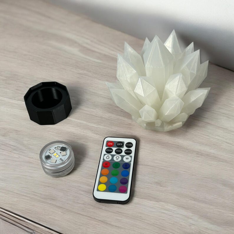 May include: A white crystal-shaped LED light with a remote control, a black cylindrical holder, and a small silver LED light. The remote has color options. The items are on a light wood-grain surface.