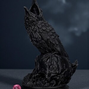 May include: A black raven statue with an open beak, perched on a textured base. The raven is detailed with visible feathers. A pink die with the number 2 is in the foreground. The background is a dark blue.