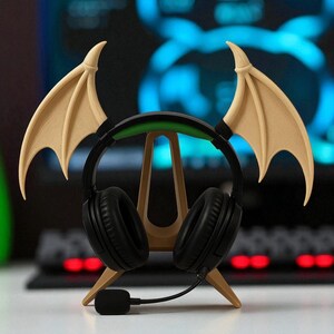 May include: Black gaming headphones with a microphone, resting on a wooden stand shaped like a bat. The stand has two beige wings attached to the top. The headphones have a green band and are on a white surface.