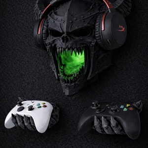 May include: A black skull-shaped wall decor with glowing green eyes and a headset. Two game controllers, one white and one black, are positioned below. The skull has horns and is wearing a headset with red accents.
