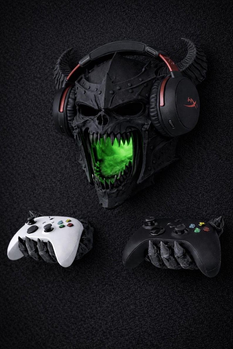 May include: A black skull-shaped wall decor with glowing green eyes and a headset. Two game controllers, one white and one black, are positioned below. The skull has horns and is wearing a headset with red accents.