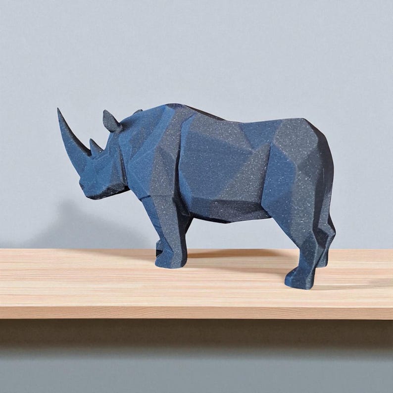 May include: A dark gray geometric rhino sculpture with a prominent horn. The rhino is standing on a light brown wooden shelf against a gray wall. The sculpture has a faceted design, giving it a modern look.