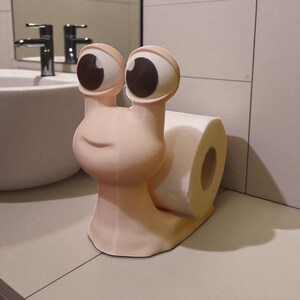 May include: A whimsical, light pink snail-shaped toilet paper holder. The holder features large, cartoonish eyes with brown pupils and a smiling face. A roll of white toilet paper is held in place by the snail's body, adding a touch of fun to bathroom decor.