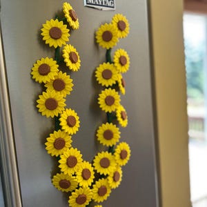 May include: A collection of yellow sunflower magnets with brown centers and green stems, arranged in a decorative pattern on a stainless steel refrigerator. The magnets are clustered together, creating a floral display.