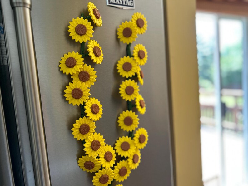 May include: A collection of yellow sunflower magnets with brown centers and green stems, arranged in a decorative pattern on a stainless steel refrigerator. The magnets are clustered together, creating a floral display.