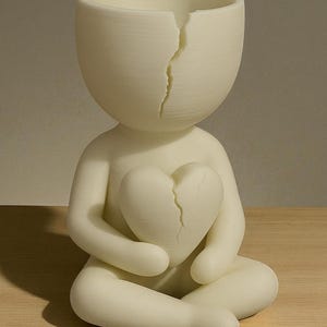 May include: A cream-colored figurine of a person seated with legs crossed, holding a heart. Both the head and the heart have a crack running down the middle. The figurine is made of a smooth material.