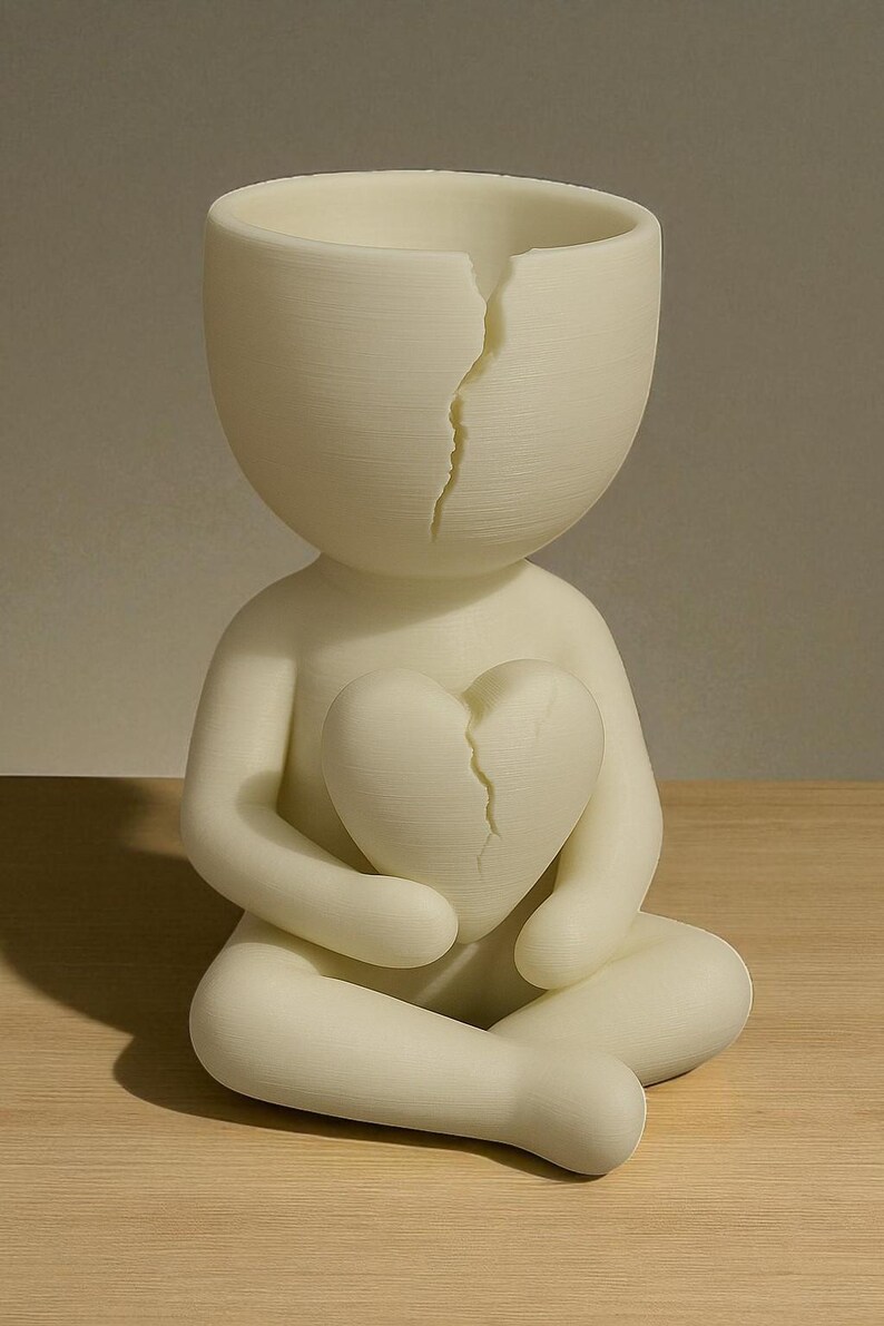 May include: A cream-colored figurine of a person seated with legs crossed, holding a heart. Both the head and the heart have a crack running down the middle. The figurine is made of a smooth material.