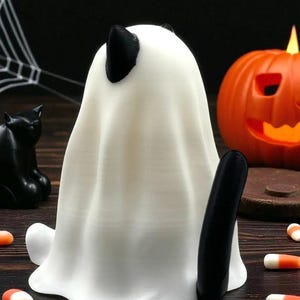 May include: A white ghost figurine with black ears and tail, a black cat figurine, and an orange jack-o'-lantern. Candy corn is scattered around the base of the ghost. Halloween-themed decorations.