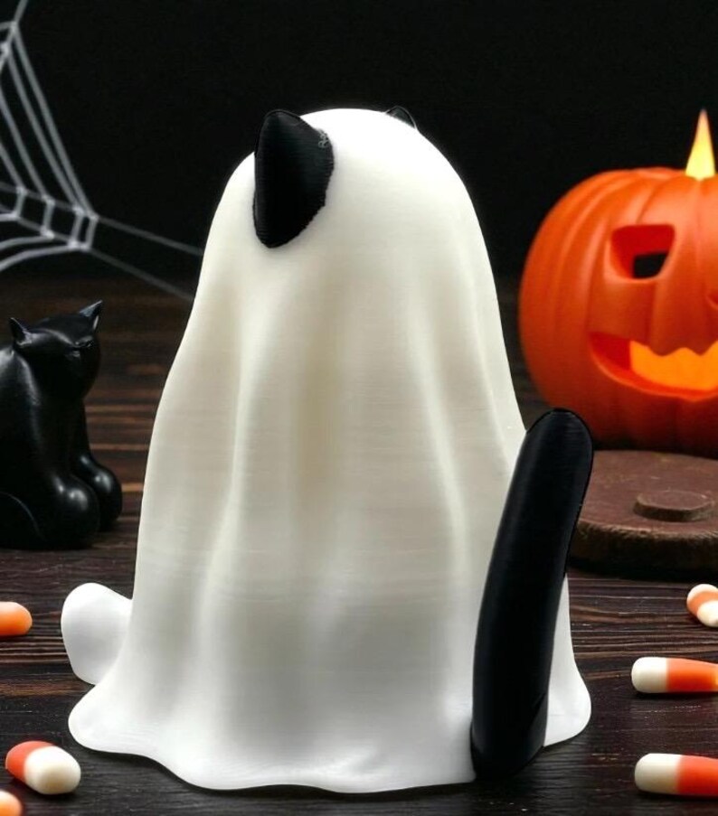 May include: A white ghost figurine with black ears and tail, a black cat figurine, and an orange jack-o'-lantern. Candy corn is scattered around the base of the ghost. Halloween-themed decorations.
