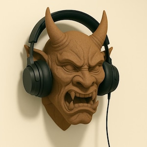 May include: A brown, textured wall-mounted headphone holder shaped like a demonic face with horns and fangs. Black headphones are placed on the holder, with the cord hanging down. The holder is mounted on a cream-colored wall.