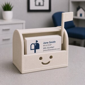 May include: A cream-coloured business card holder in the shape of a mailbox, featuring a smiling face design. It holds business cards printed with "Jane Smith, REALTOR" and contact details. The mailbox has a small flag.