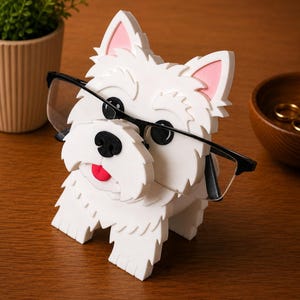 May include: A white dog-shaped eyeglass holder with black-framed glasses. The holder has pink inner ears, a black nose, and a red tongue. The holder is made of a textured material and sits on a wooden surface.
