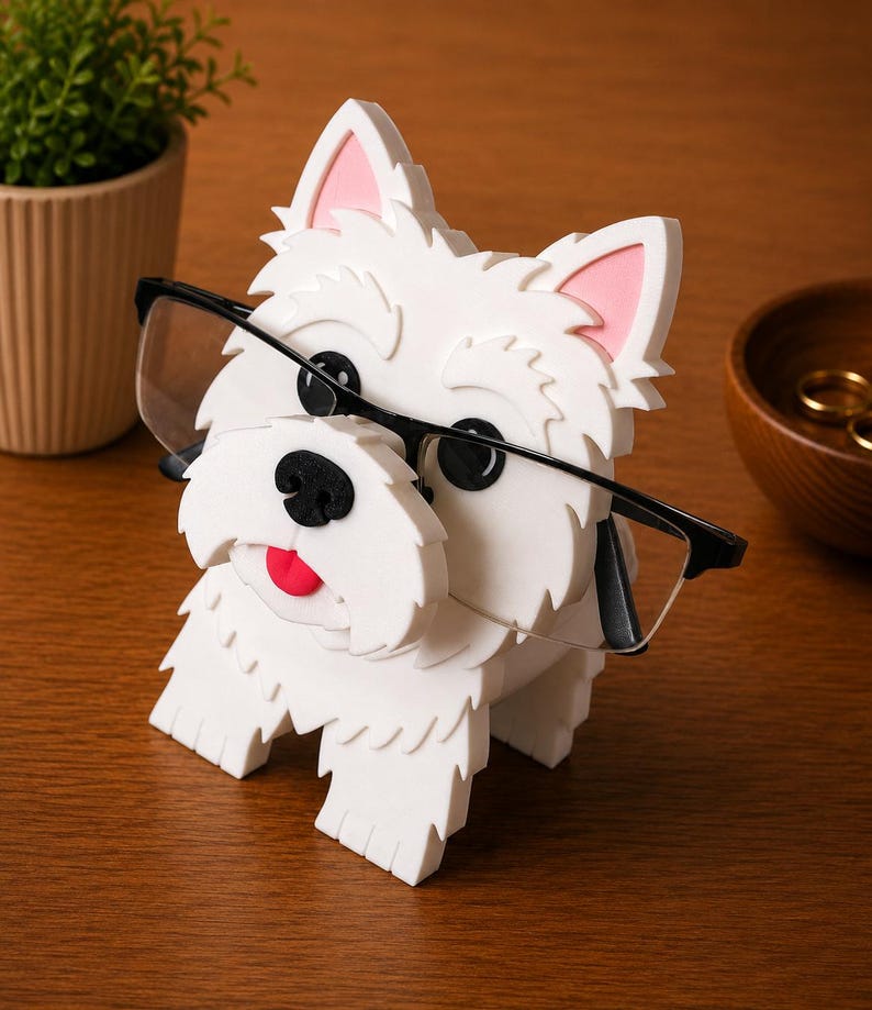 May include: A white dog-shaped eyeglass holder with black-framed glasses. The holder has pink inner ears, a black nose, and a red tongue. The holder is made of a textured material and sits on a wooden surface.
