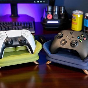 May include: Two gaming console controllers, one white and one black, rest on miniature sofa-shaped stands. The stands are upholstered in green and blue fabric, with wooden legs. A keyboard, monitor, and other gaming accessories are in the background.