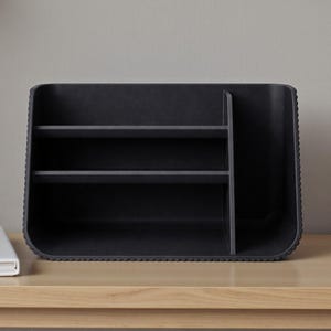 May include: A black desk organizer with two shelves and a vertical divider. The organizer has a textured, ridged edge and is made of a matte material. It sits on a light-colored wooden surface.