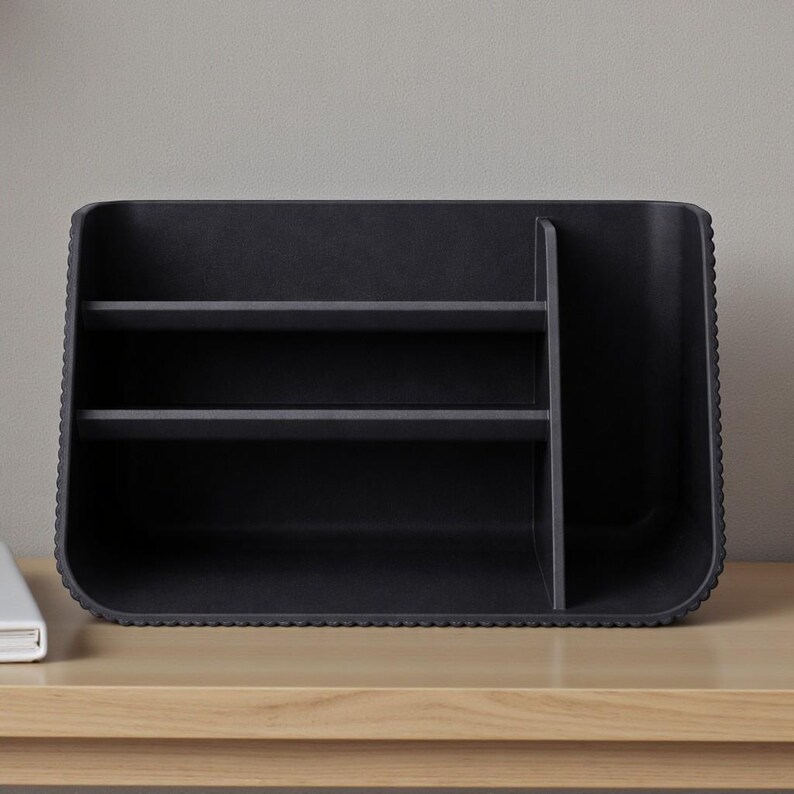 May include: A black desk organizer with two shelves and a vertical divider. The organizer has a textured, ridged edge and is made of a matte material. It sits on a light-colored wooden surface.