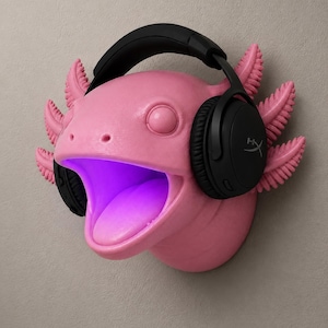 May include: A pink axolotl wall mount with black headphones. The axolotl has a wide open mouth with a purple glow inside. The headphones have the HyperX logo on the earcups. The axolotl has feathery gills.