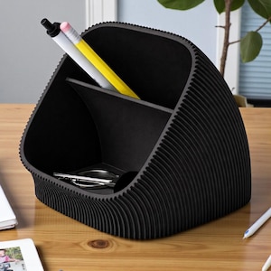 May include: A black desk tidy with a distinctive, ridged design. The organiser has several compartments, holding pens, pencils, and small office supplies. The desk tidy is made of a solid black material and sits on a wooden desk.