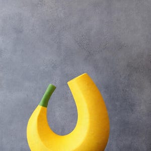 May include: A modern, abstract yellow vase with a green stem. The vase has a curved, banana-like shape with a textured surface. The background is a solid gray.
