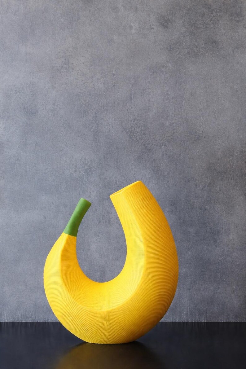 May include: A modern, abstract yellow vase with a green stem. The vase has a curved, banana-like shape with a textured surface. The background is a solid gray.
