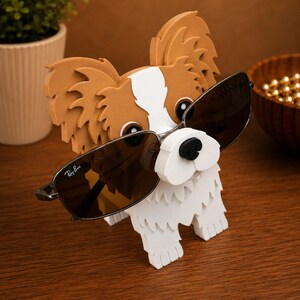 May include: A decorative wooden dog-shaped sunglasses holder. The dog is white with brown and tan accents and is wearing Ray-Ban sunglasses. The holder is on a wooden surface.