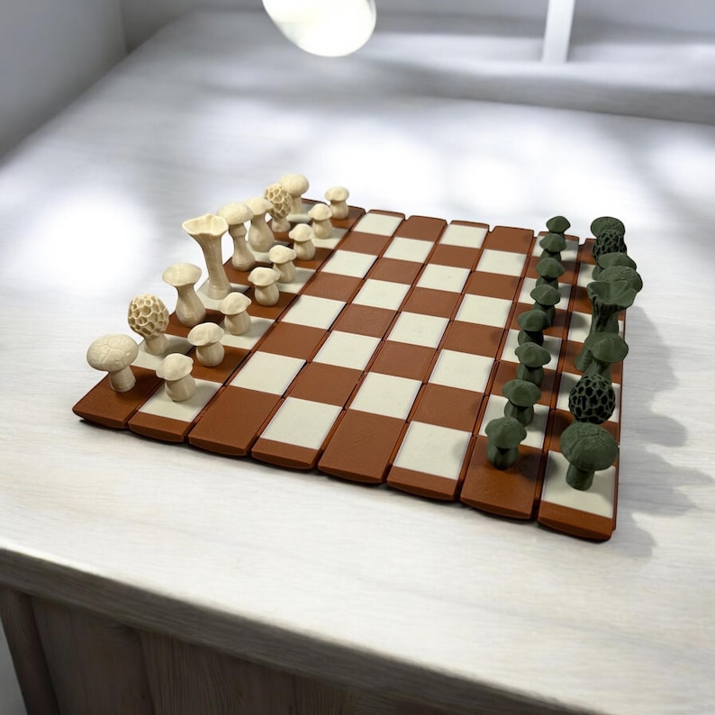 May include: A novelty chess set with mushroom-shaped pieces in cream and green. The board has a brown and cream checkered pattern. The pieces are detailed, adding a whimsical touch to the classic game. The set is displayed on a light-coloured surface.