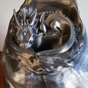 K&ouml;nnte beinhalten: Silberfarbene Skulptur eines Babydrachens, der sich in einer kokonartigen Struktur zusammenrollt. Der Drache hat detaillierte Merkmale, darunter H&ouml;rner und Schuppen. Der Kokon hat ein strukturiertes, drapiertes Aussehen.