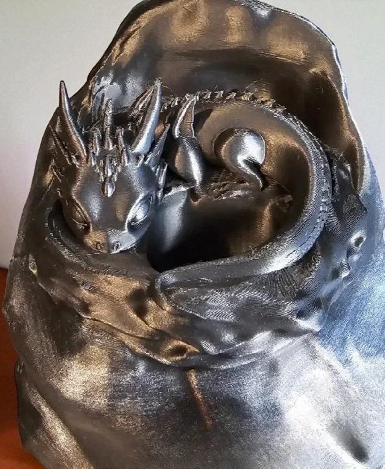 K&ouml;nnte beinhalten: Silberfarbene Skulptur eines Babydrachens, der sich in einer kokonartigen Struktur zusammenrollt. Der Drache hat detaillierte Merkmale, darunter H&ouml;rner und Schuppen. Der Kokon hat ein strukturiertes, drapiertes Aussehen.