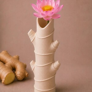 Op de afbeelding: Een beige keramische vaas in de vorm van een gemberwortel, met een roze lotusbloem. De vaas heeft een textuur en decoratieve details. De bloem heeft roze bloemblaadjes en een gele kern.