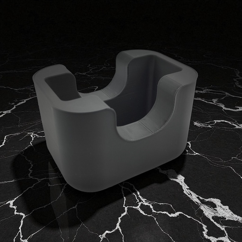 May include: A dark gray, modern-style desk organizer with a curved design. The organizer has a square base and a rounded top with two open sections. The object is made of a smooth material and sits on a black and white marbled surface.