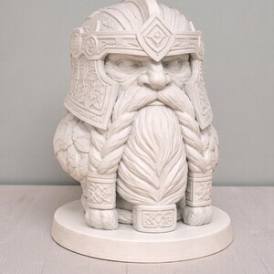 May include: An off-white sculpture of a dwarf with a detailed helmet and braided beard. The sculpture is mounted on a round base. The helmet and beard feature intricate carvings. A decorative item.