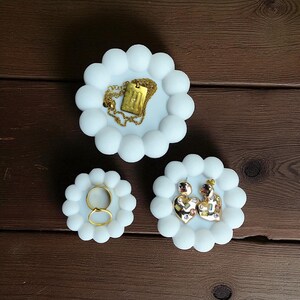 May include: Three white, cloud-shaped jewelry dishes. One holds a gold necklace with a rectangular pendant. Another holds two gold rings. The third holds a pair of heart-shaped earrings with colorful accents.
