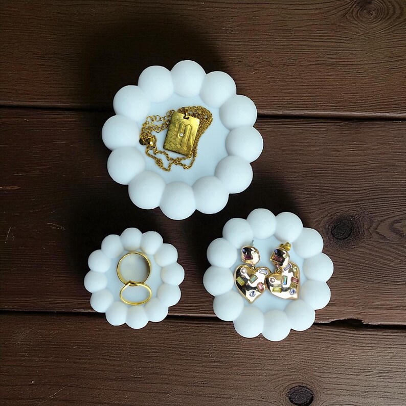 May include: Three white, cloud-shaped jewelry dishes. One holds a gold necklace with a rectangular pendant. Another holds two gold rings. The third holds a pair of heart-shaped earrings with colorful accents.